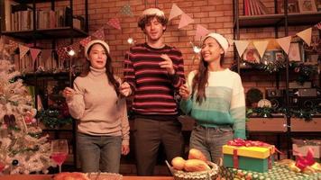 Group of three young friends happiness dancing and joyful swaying sparkler fireworks, celebrate Christmas festival at dining table with gifts and foods, home decorated for New Year's party night. video