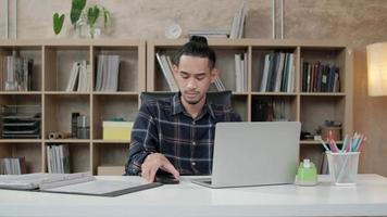 Young male worker of Asian ethnicity, laptop to do creative work, mobile phone communication on desk, bookshelf behind at casual workplace, startup business person, and online e-commerce occupation.