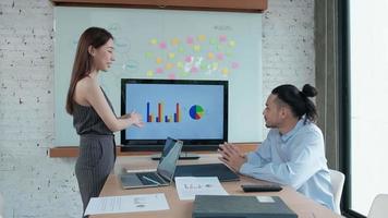 Two young and smart Asian co-workers are brainstorming at office. Female colleague presents business chart to male partner using a laptop in a meeting room, discussing the company's finances project.