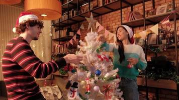 Zwei junge Paare sind zusammen verliebt, fröhlich und schmücken glücklich einen weißen Weihnachtsbaum mit Ornamenten im Wohnzimmer des Hauses, bereiten Sie sich auf eine Feier zum Neujahrsfest vor.