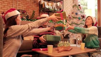 Sorprenda a tres amigos y familiares dándoles regalos juntos e intercambiándolos mientras las felices vacaciones en la sala de estar de su casa están decoradas para la celebración del festival navideño y la fiesta de año nuevo. video