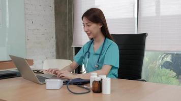 Young beautiful Asian female doctor with stethoscope and uniform working and video chatting with a smile about healthy with laptop computer on a desk, medicine nearby at hospital clinic office.