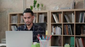 Young male worker of Asian ethnicity, laptop to do creative work, mobile phone communication on desk, bookshelf behind at casual workplace, startup business person, and online e-commerce occupation. video