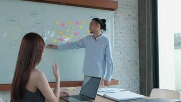 Two colleagues and coworkers of Asian ethnicity brainstorm and meeting finance project discuss with business plan in conference room with colorful sticky notes paper on writing board in the office. video