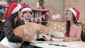 drie arbeidersteam met rode hoeden en hond, vrienden en collega's die praten en ontspannen op kantoor voor bedrijfsvakanties, feestelijk versieren voor het vieren van kerstfeest en nieuwjaarsdag.