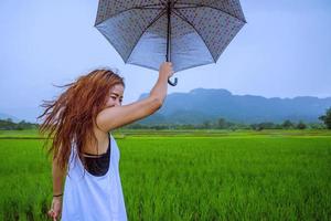 las mujeres asiáticas viajan relajarse en las vacaciones. la mujer de pie sostiene un paraguas bajo la lluvia feliz y disfrutando de la lluvia que cae. viajando en campiña, campos de arroz verde, viaje a tailandia. foto