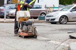 A worker cuts the old asphalt on the road with a gasoline cutter against the background of a city street and passing cars in blur. photo