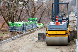 A road roller on a fenced building site with a sidewalk under construction in the background and stacks of gray paving slabs in blur. photo