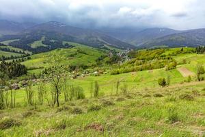 The mountain slope is covered with thick green grass against the backdrop of the picturesque landscape of the Carpathian Mountains shrouded in mist and villages in the valley. photo