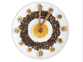 Grains of roasted coffee, mugs of dried orange and banana, cinnamon stick, anise star lie on a porcelain plate in the form of a clock photo