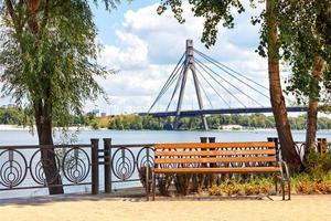 Summer landscape with a wooden bench on the Dnipro embankment against the background of the Northern bridge over the river in blur.