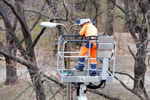 A utility worker changes a light bulb on a high concrete pole of street lighting in a city park. photo