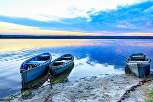 Old blue-green boats moored to the shore by old iron chains and stand on the shore of a calm river against the background of the morning horizon. photo