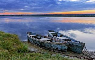 Two old dilapidated boats are moored with a metal chain to the bank of a calm river in the early morning against the backdrop of a cloudy sky and the rising sun. photo