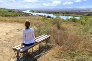 A young woman sits on a wooden bench and looks at the calm Southern Bug River on a sunny autumn day. photo