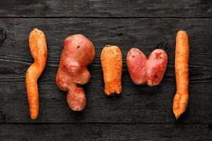 Ugly potato in the heart shape and twisted carrot on a black wooden background. Vegetable or food waste concept. Top view, close-up. photo