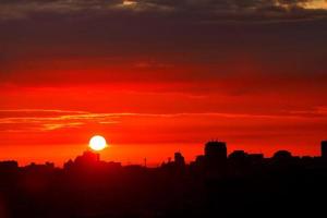 A bright disc of sun rises in the early morning over the city's silhouettes of houses, filling the sky with red light. photo