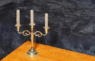 A metal gilded candlestick with three candles stands on a yellow wooden table against a dark gray velor background. photo