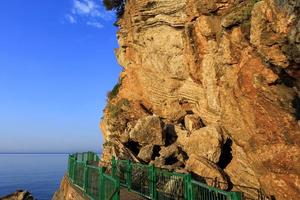 The road along the sea coast, surrounded by a green metal fence, is illuminated by the rays of the morning golden sun near a rocky cliff. photo