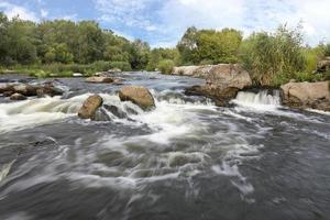 el rápido flujo del río, costas rocosas, rápidos, vegetación de color verde brillante y un cielo azul nublado en verano: una vista frontal foto