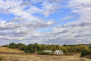 An abandoned rural house stands on the side of a rural road at the edge of a field under a cloudy sky. photo