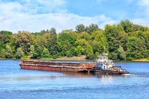 A river tug is pushing a rusty barge along the river along the shore.