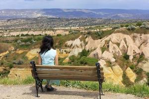 A young beautiful woman in a turquoise dress sits on a wooden bench on a hill against the blurred landscape of mountain caves in the valleys and canyons of Cappadocia
