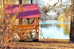 cenador de madera con una mesa y bancos de picnic al aire libre en el fondo de hojas caídas cerca de un lago del bosque. foto