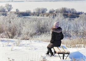 A young woman sits on a wooden bench and looks at the calm Southern Bug River on a sunny winter day.