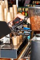 The barista prepares coffee and pours it into a paper cup against the backdrop of a coffee maker and a slide of empty paper cups. photo