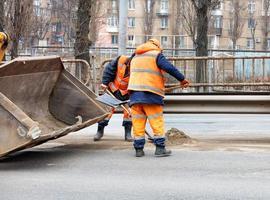 The road workers use shovels to scrape off the sand accumulated between the lanes of the road and load it into the grader bucket. photo