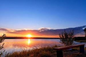 el brillante sol naciente se eleva sobre el horizonte detrás de un ancho río y se refleja en una superficie ondulada del agua. foto