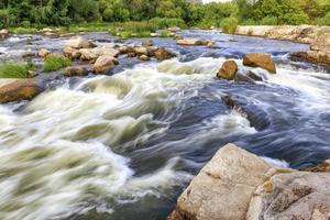 The rapid flow of the river in the blur, rocky shores, boulders and rapids, bright green vegetation on the other side of the shore. photo