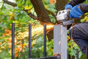 Craftsman cuts metal pillar metal with disc angle grinder on blurred background of green park. photo