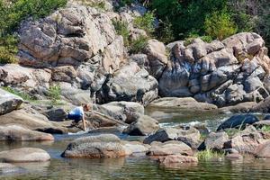 A young lady dips her hands into the foamy streams of water on stone boulders on the bank of a forest river on a sunny summer day. photo