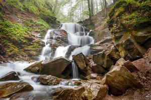 The rapid flow of mountain river water cascades and whitish threads makes its way through the rocky forest hilly terrain in the spring. photo