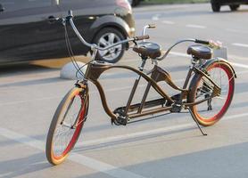 A tandem bicycle with scarlet wheel rims is parked in a parking lot in the evening sun. photo