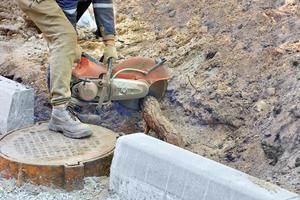 A worker using a portable gasoline saw clears the site and cuts the powerful roots of a tree to equip a manhole. photo