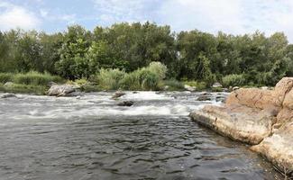 Fast river flow, rocky shores, rapids, bright green vegetation and a cloudy blue sky in summer photo