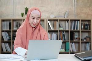 Beautiful businesswoman of Asian ethnicity works on e-commerce using laptop, internet communication in small business office. Attractive person, traditionally Islam dresses by wearing hijab. photo