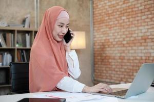 Beautiful businesswoman who Islamic female person is talking on her mobile phone communication and happy smiling because internet online order in laptop, modern lifestyle, e-commerce occupation. photo