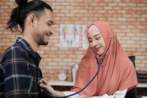 Young beautiful woman doctor is health examining male patient in office of hospital clinic and advising with a smile on medicines. This Asian medical specialist is an Islamic person wearing a hijab. photo