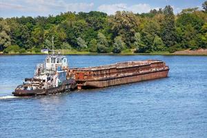 A river tugboat with an empty rusty barge goes crosses a wide river.