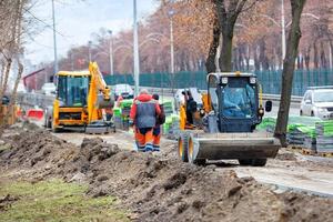 una niveladora de carreteras compacta se mueve a lo largo de una acera en construcción frente a un sitio de construcción y un equipo de trabajadores de la carretera en el desenfoque. foto