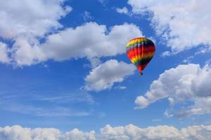 un abigarrado globo de aire caliente multicolor levanta una canasta con turistas en el cielo azul entre nubes blancas.
