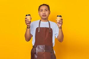 Portrait of confused handsome man holding two cups of coffee and looking ahead isolated on yellow background photo