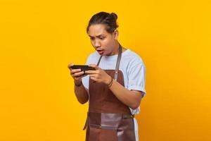 Portrait of handsome asian man wearing apron looking annoyed while playing game on smartphone photo