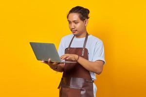 Portrait of handsome young barista using laptop looking serious doing office work on yellow background photo