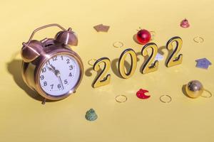 Alarm clock, Christmas decor and numbers 2022 with hard shadow on yellow background. New year celebration minimal concept. Close up, copy space photo