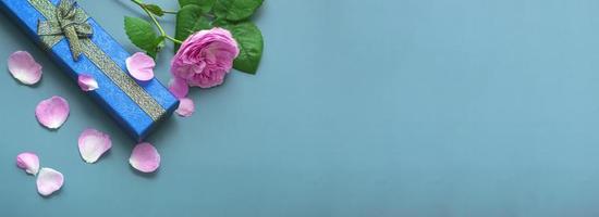 Top view for Father's Day on blue background with rose petals. Close up of gift blue box with gold bows and pink rose. Banner photo